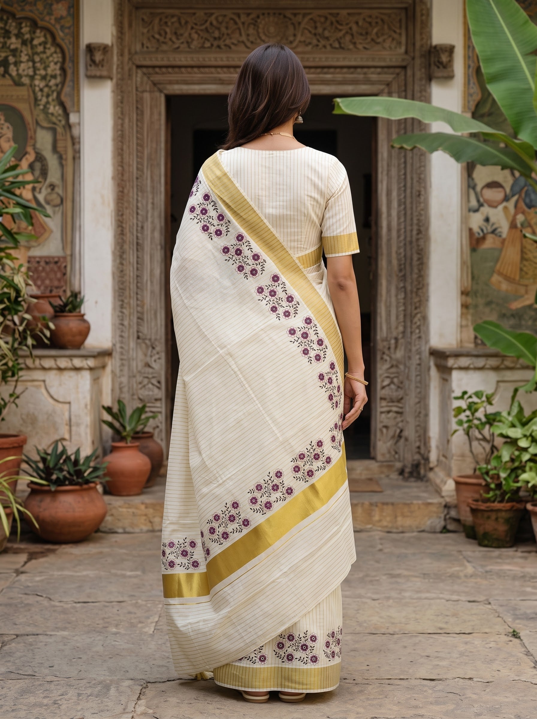 A woman viewed from behind wearing a cream-colored saree with gold borders and purple floral patterns, standing in front of an ornate stone doorway with potted plants.