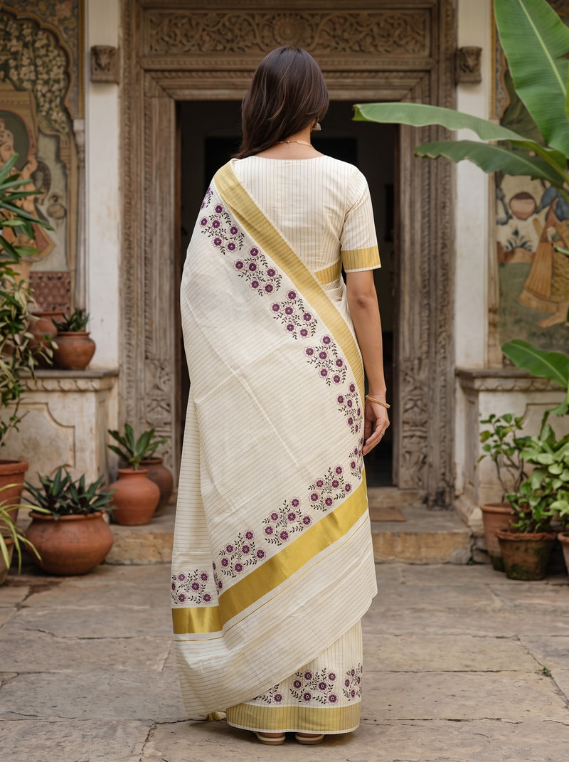 A woman viewed from behind wearing a cream-colored saree with gold borders and purple floral patterns, standing in front of an ornate stone doorway with potted plants.