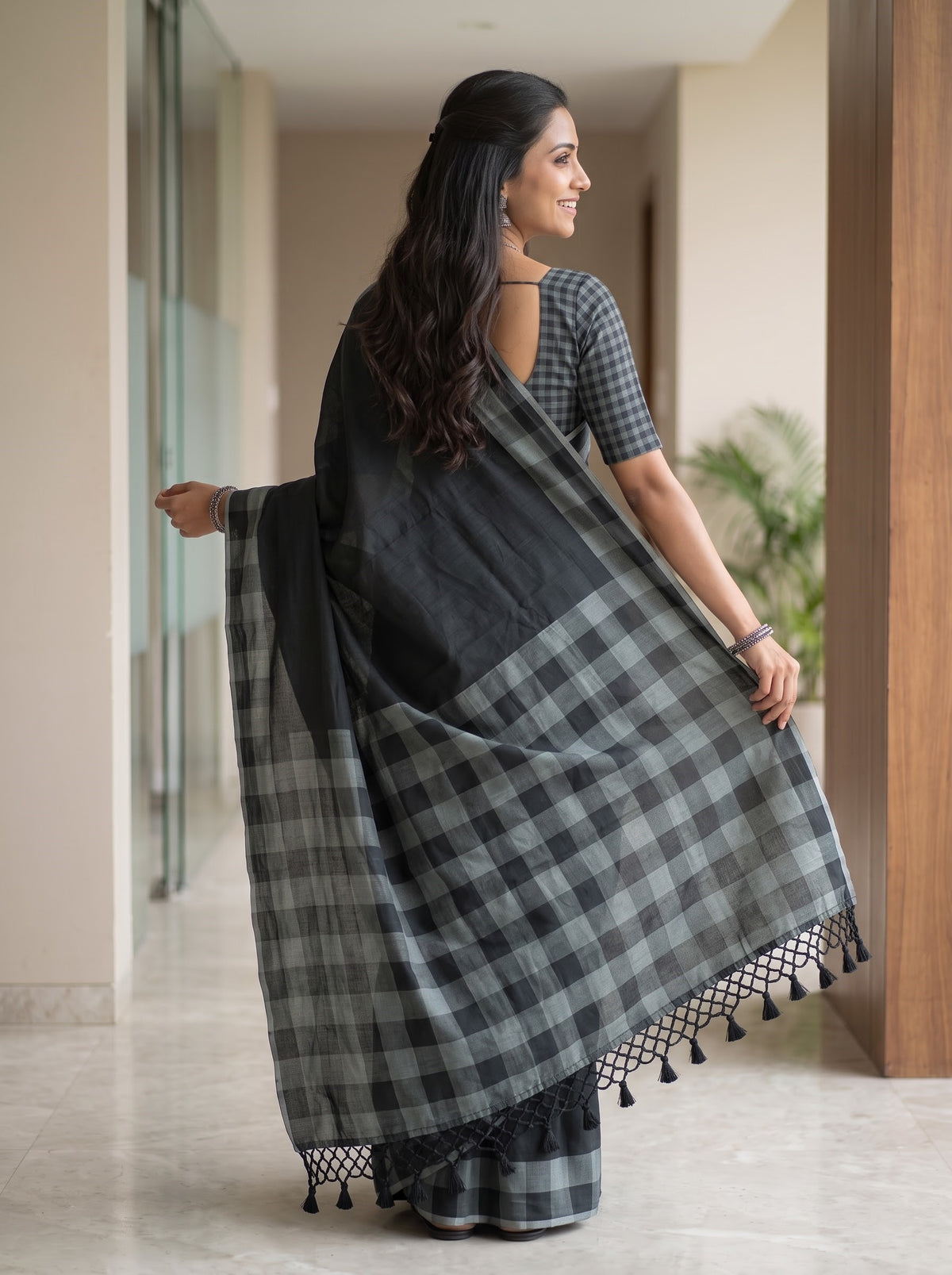 A woman wearing a black and gray checkered saree with a matching checkered blouse, standing in a modern indoor hallway and holding out the saree drape.