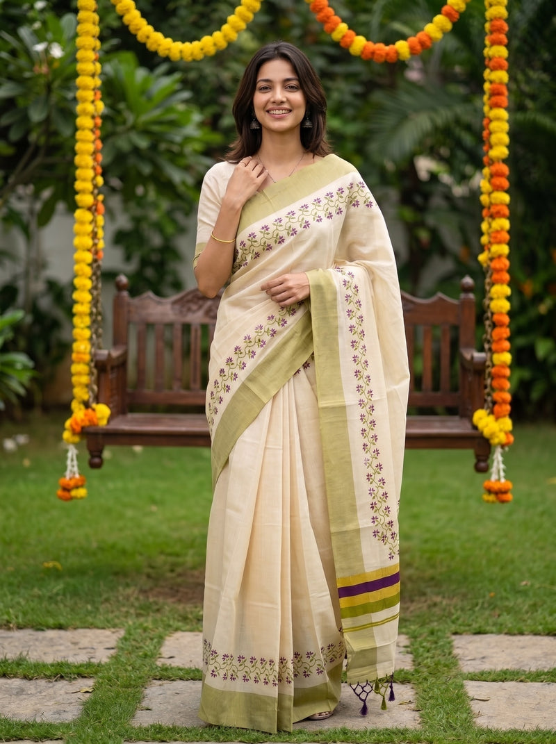 A woman wearing a cream-colored cotton saree with olive green borders and floral prints, standing in a garden decorated with marigold garlands.