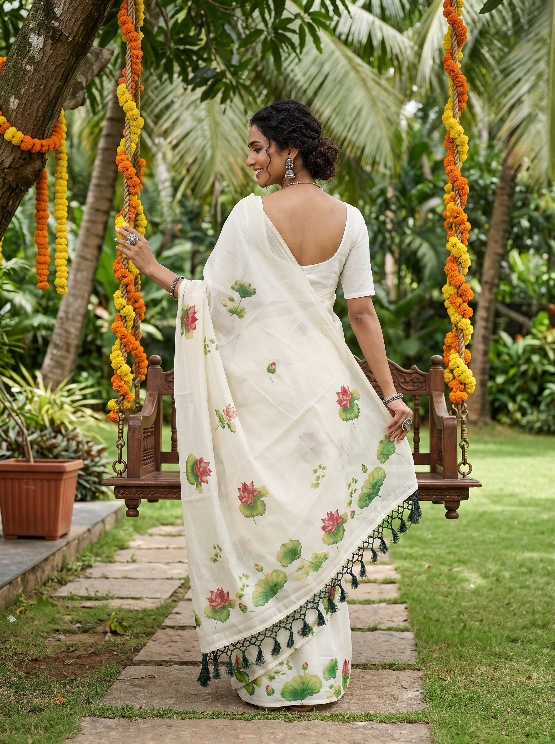 A woman wearing a white cotton saree with green lotus and pink lotus floral prints, standing on a stone path in a garden next to a wooden swing decorated with orange marigold garlands.