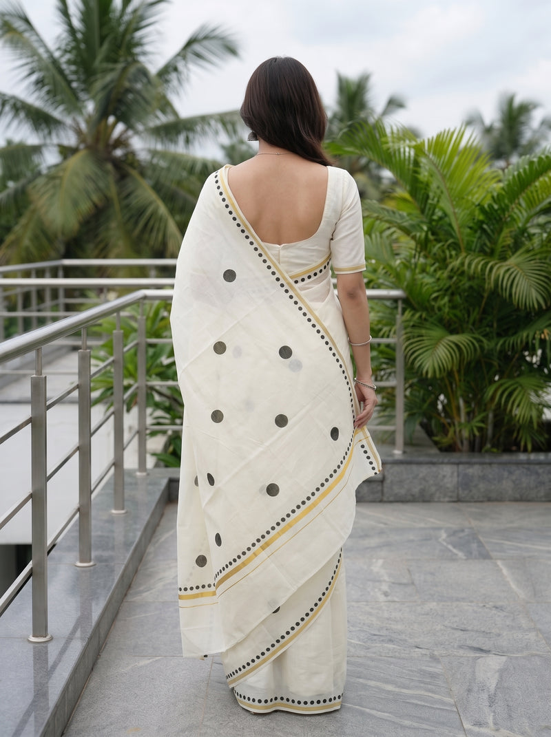 A woman viewed from behind wearing a cream-colored cotton saree with black polka dots and yellow/gold border details, standing on an outdoor balcony with palm trees in the background.