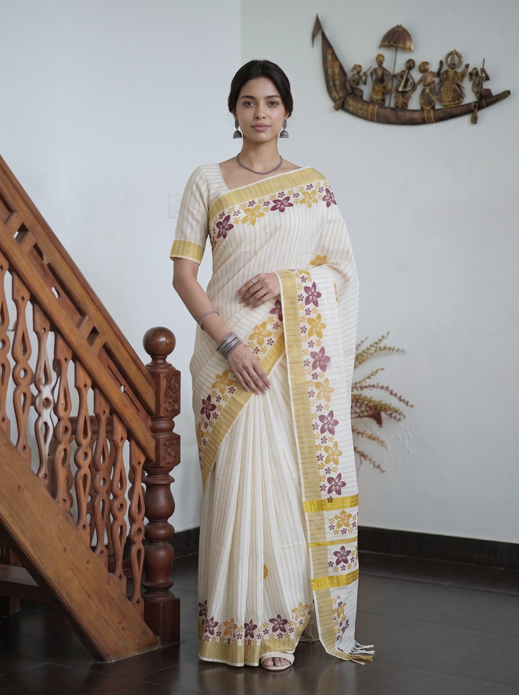 A woman wearing a cream-colored cotton saree with yellow and purple floral embroidery, standing in a home interior next to a wooden staircase.