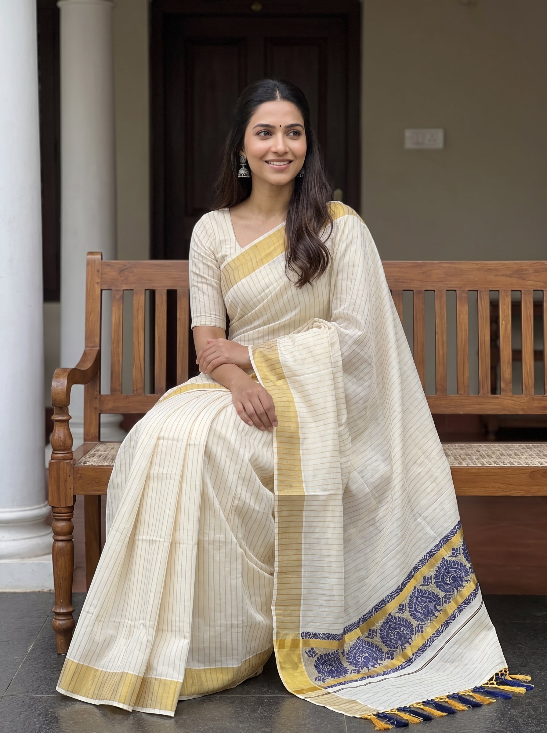 A woman wearing a cream-colored cotton saree with yellow and blue border details, sitting on a wooden bench in front of a house entrance.