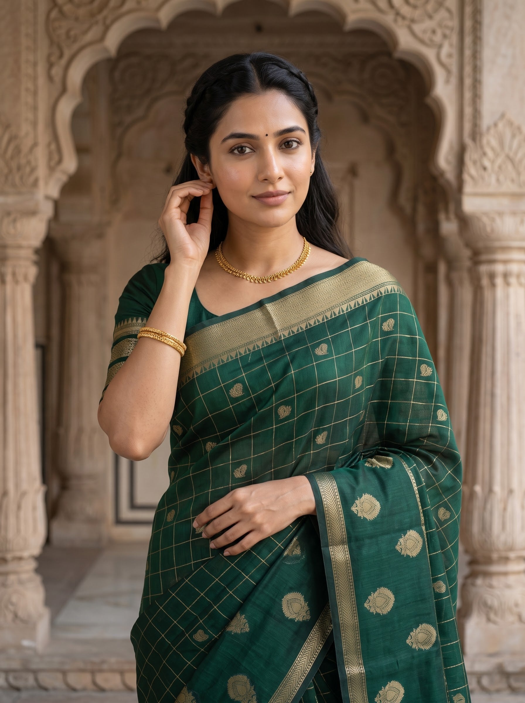 A woman wearing a dark green silk saree with gold zari checks and motifs, standing in front of a carved stone archway.