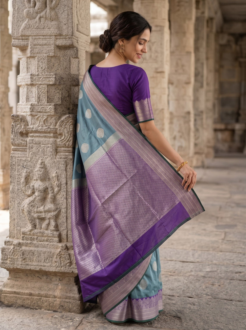 A woman wearing a purple and teal silk saree with gold zari embroidery, standing in a stone corridor.