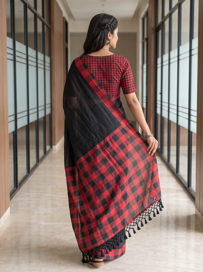 A woman viewed from behind wearing a red and black checkered saree with a solid black upper drape, standing in a modern hallway with glass-paneled walls.