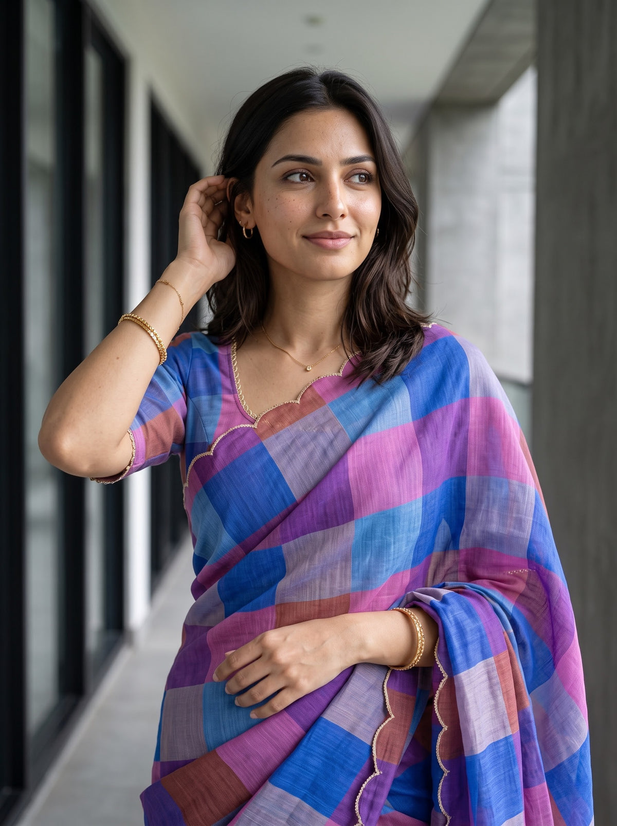A woman wearing a purple and blue checkered saree with gold jewelry, standing in a modern corridor.
