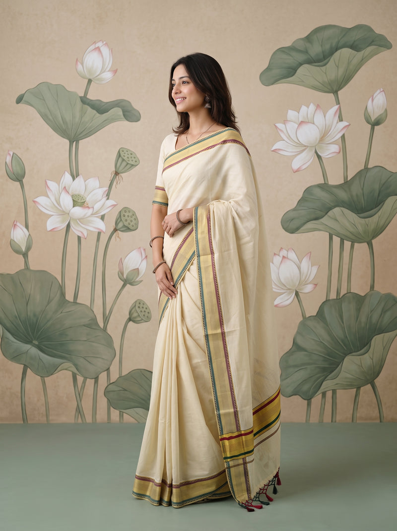 A woman wearing a cream-colored cotton saree with green, red, and gold stripes, standing in front of a beige wall decorated with lotus leaves and flowers.
