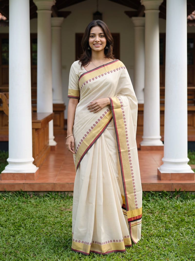 A woman wearing a cream-colored cotton saree with a gold and maroon border, standing outdoors in front of a traditional building with white pillars and green grass.