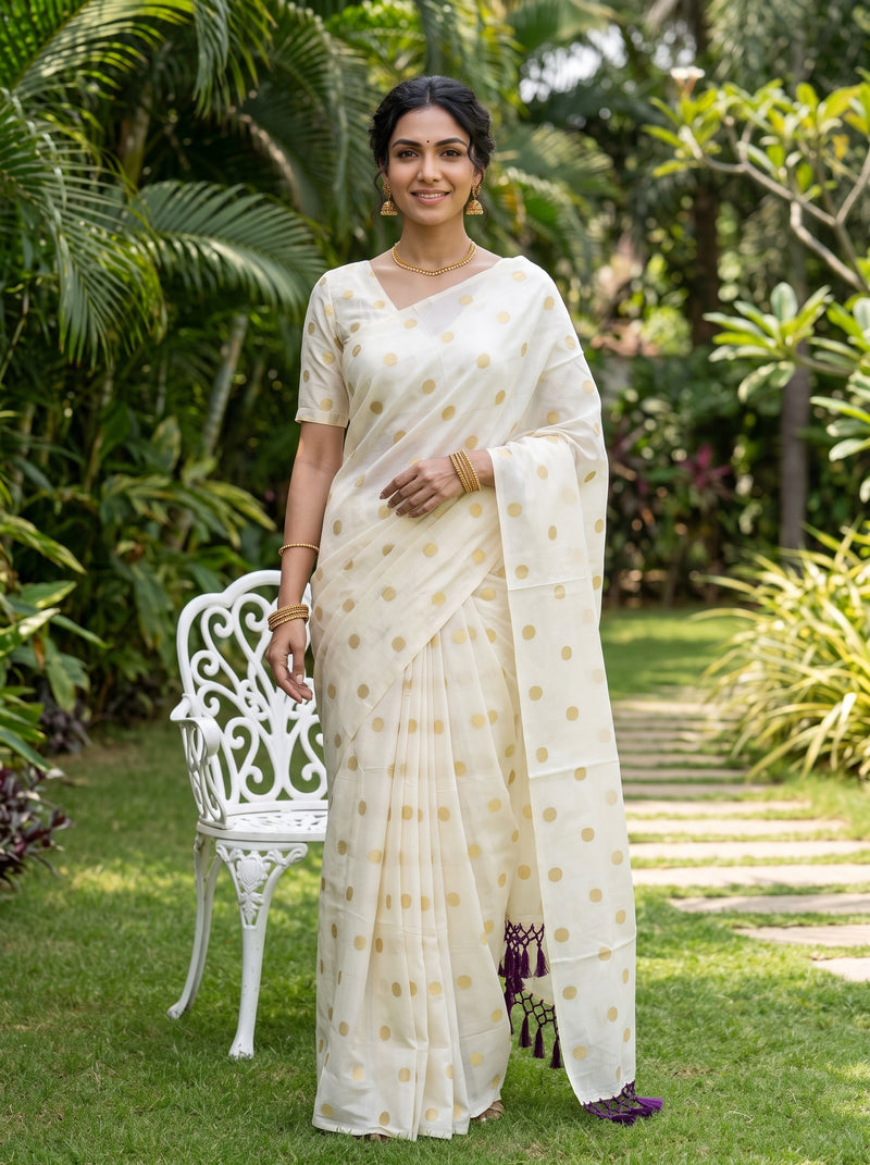 A woman wearing a cream-colored saree with gold polka dots and a matching blouse stands in a lush green garden next to a white garden chair.