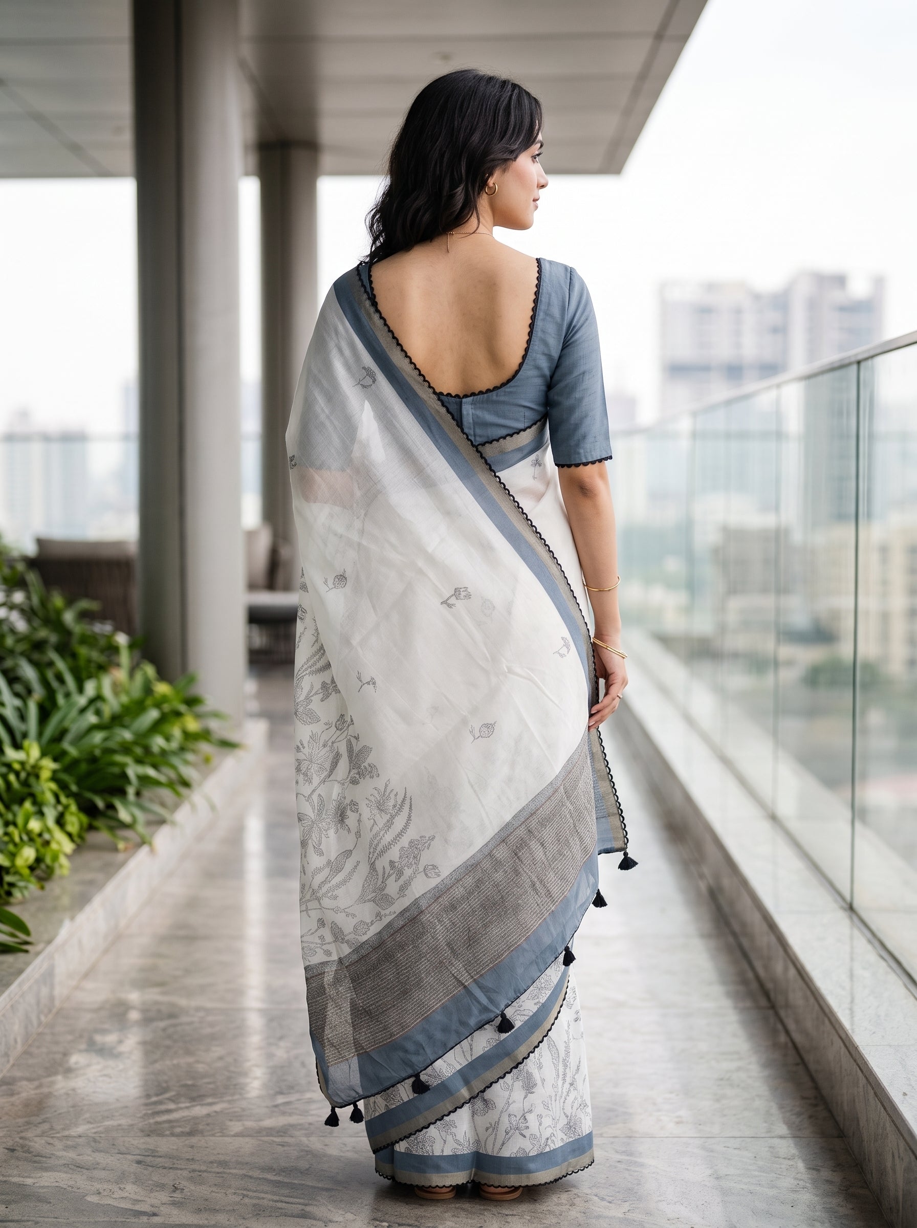 A woman stands on a modern balcony wearing a white and gray patterned saree with a slate blue blouse, viewed from behind to show the back of the garment.