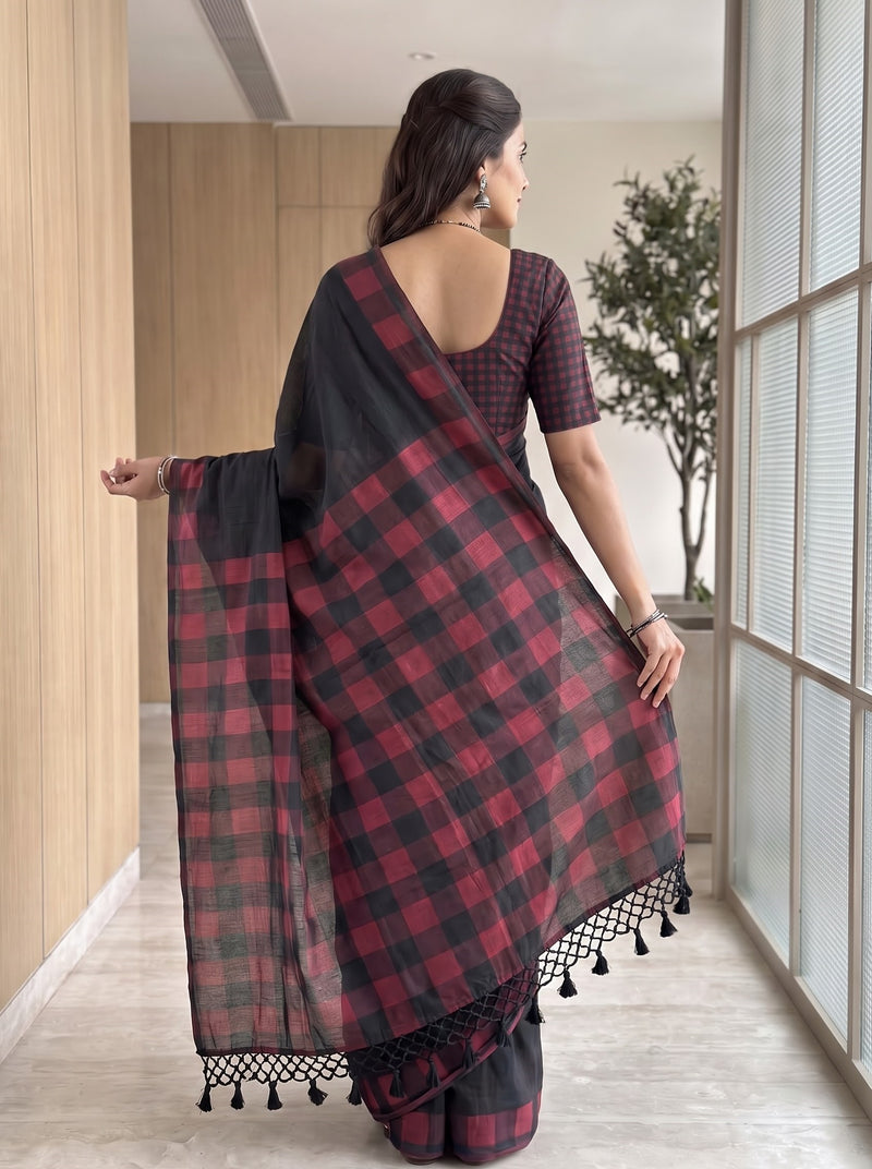 A woman wearing a black and red checkered saree with a matching checkered blouse, standing in a modern indoor hallway with wood-paneled walls and large windows.