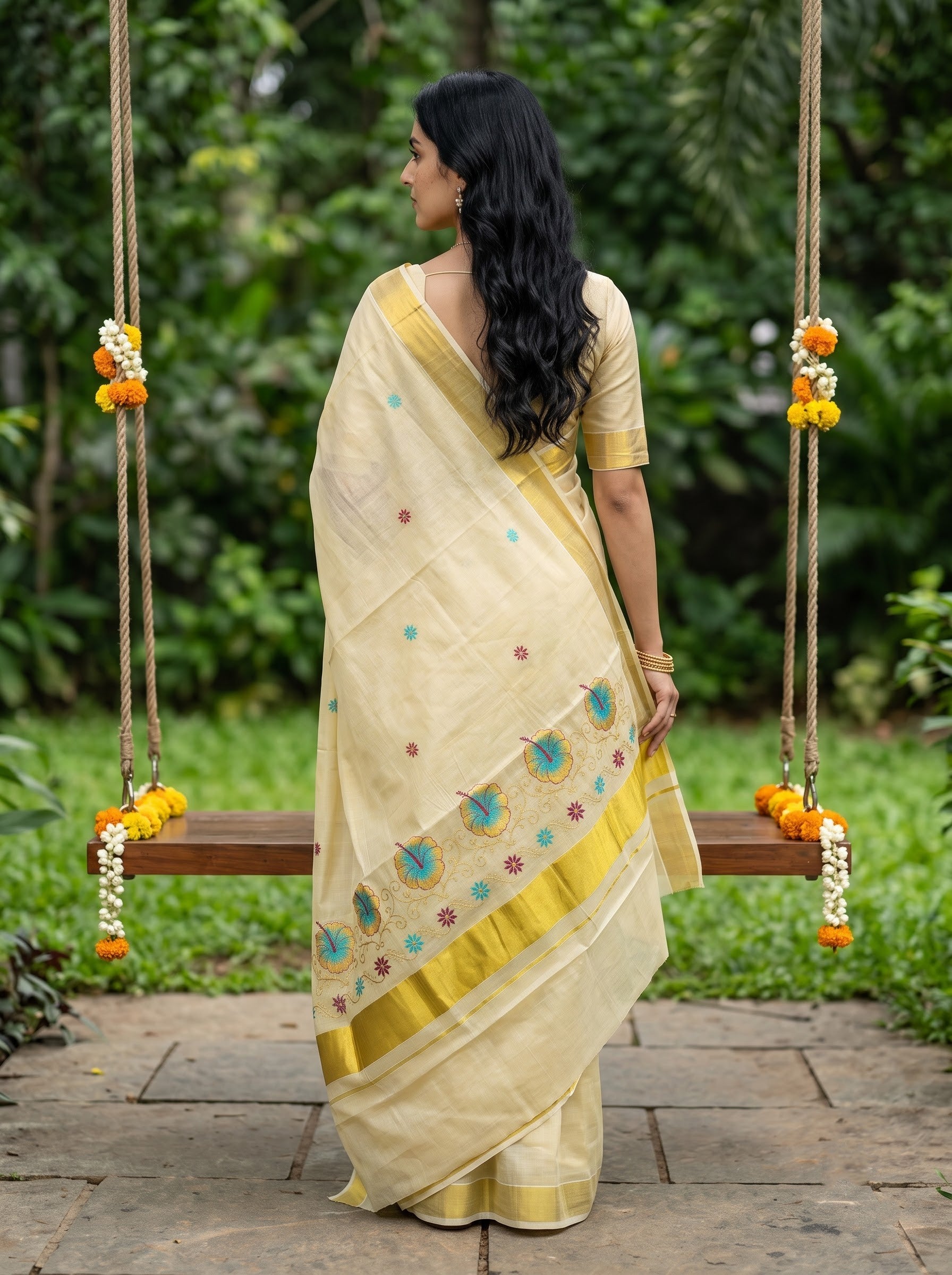 A woman wearing a cream-colored saree with gold borders and blue floral motifs, standing on a wooden swing in a lush green garden.