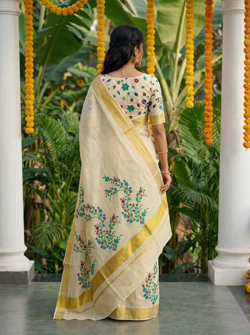 A woman viewed from behind wearing a cream-colored saree with green and red floral embroidery and a yellow border, standing in front of a backdrop of green tropical plants and hanging yellow marigold garlands.