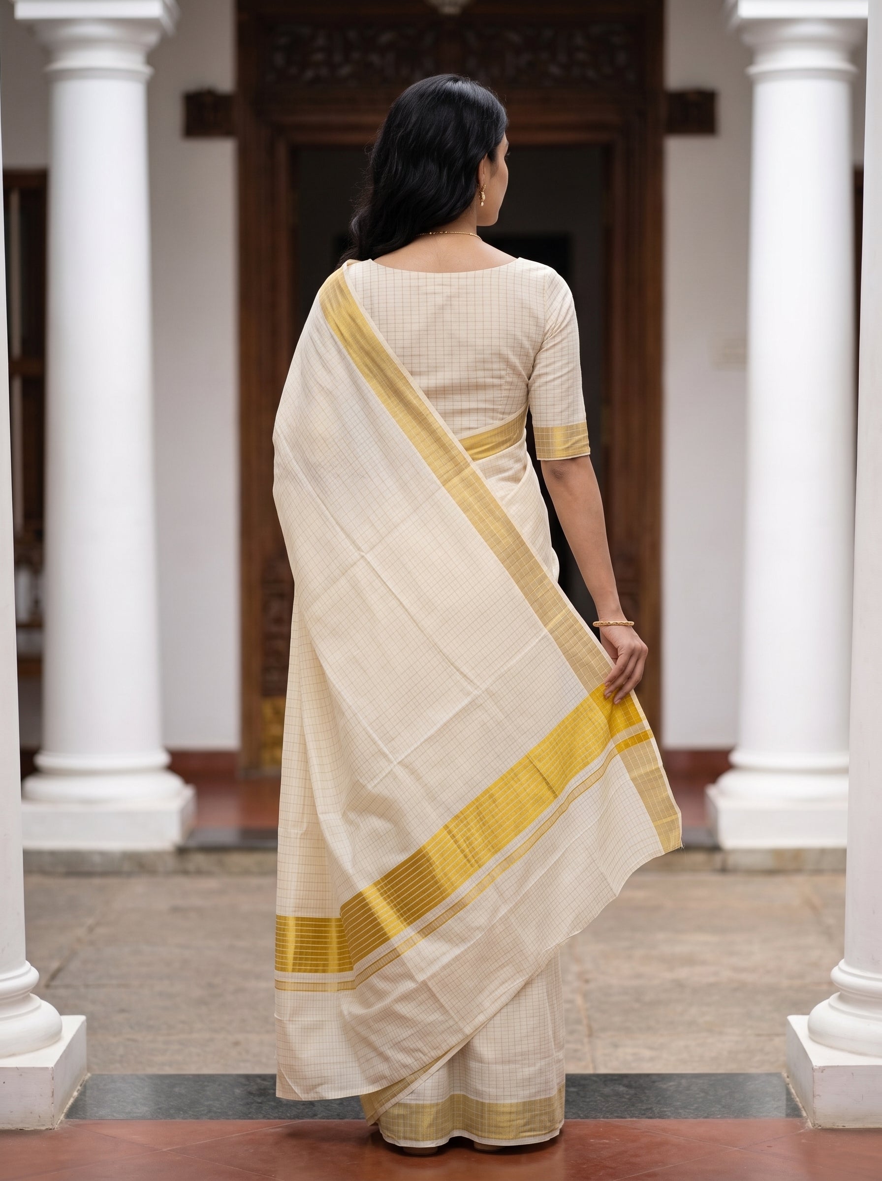 A woman viewed from behind wearing a cream-colored saree with a yellow border, standing in a corridor with white columns.