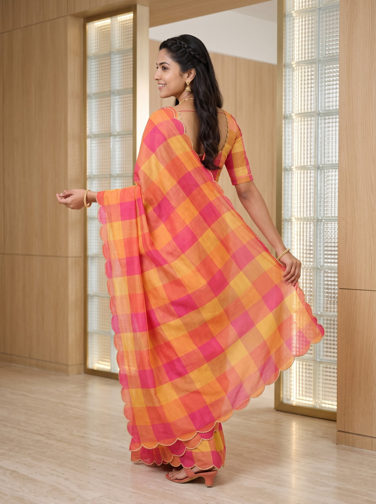 A woman wearing a bright pink and orange checkered saree with a matching blouse, standing in a modern indoor setting with light wood paneling and glass doors.