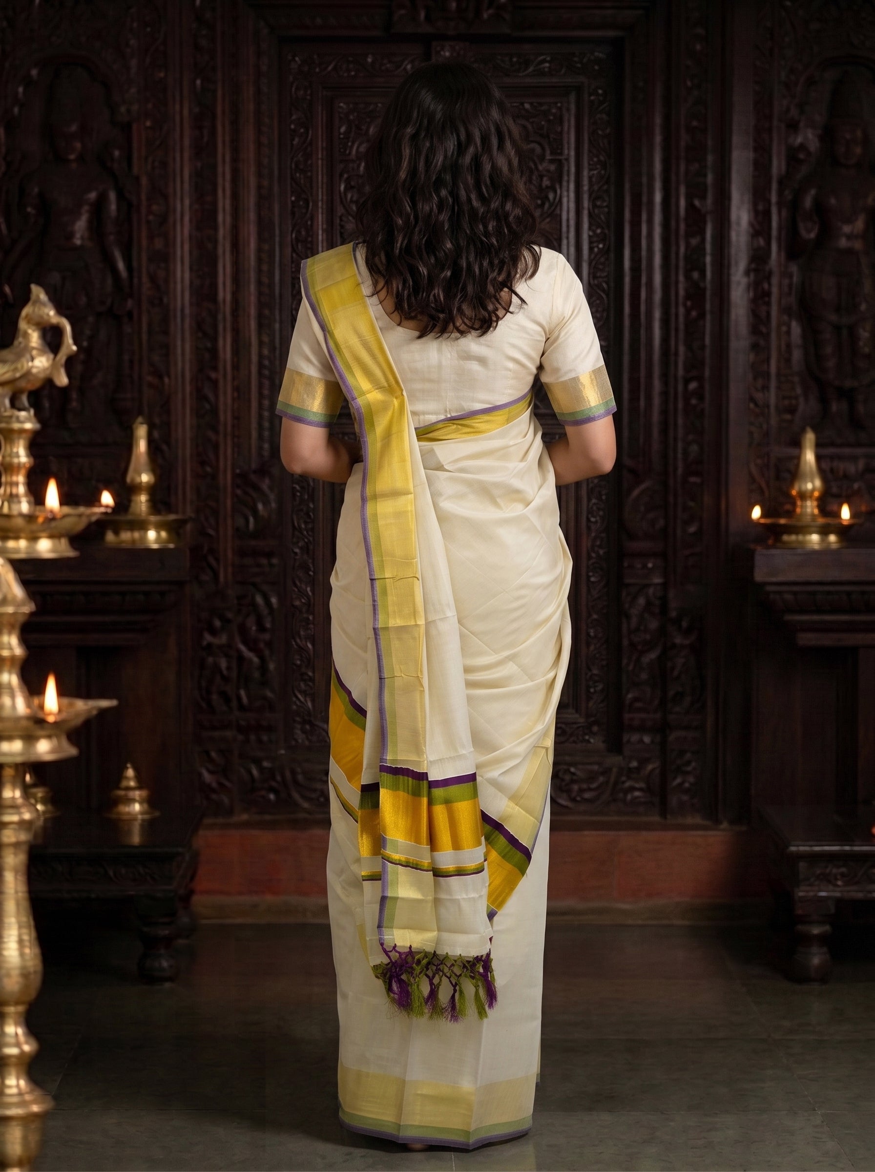 A woman viewed from behind wearing a cream-colored saree with yellow and purple border accents, standing in front of a dark carved wooden door with brass lamps on either side.