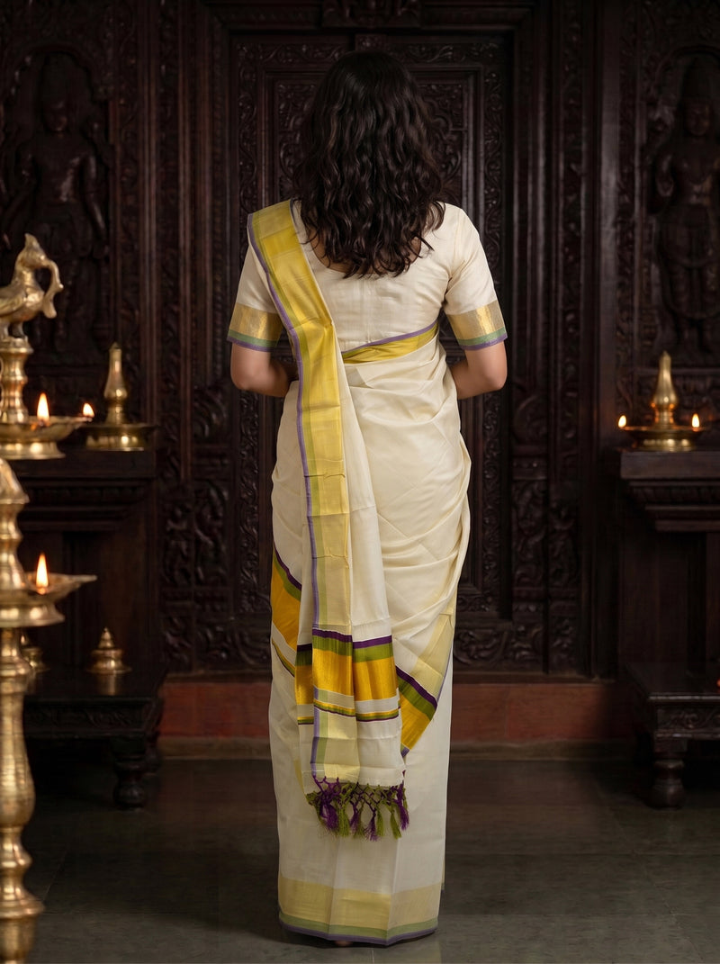 A woman viewed from behind wearing a cream-colored saree with yellow and purple border accents, standing in front of a dark carved wooden door with brass lamps on either side.