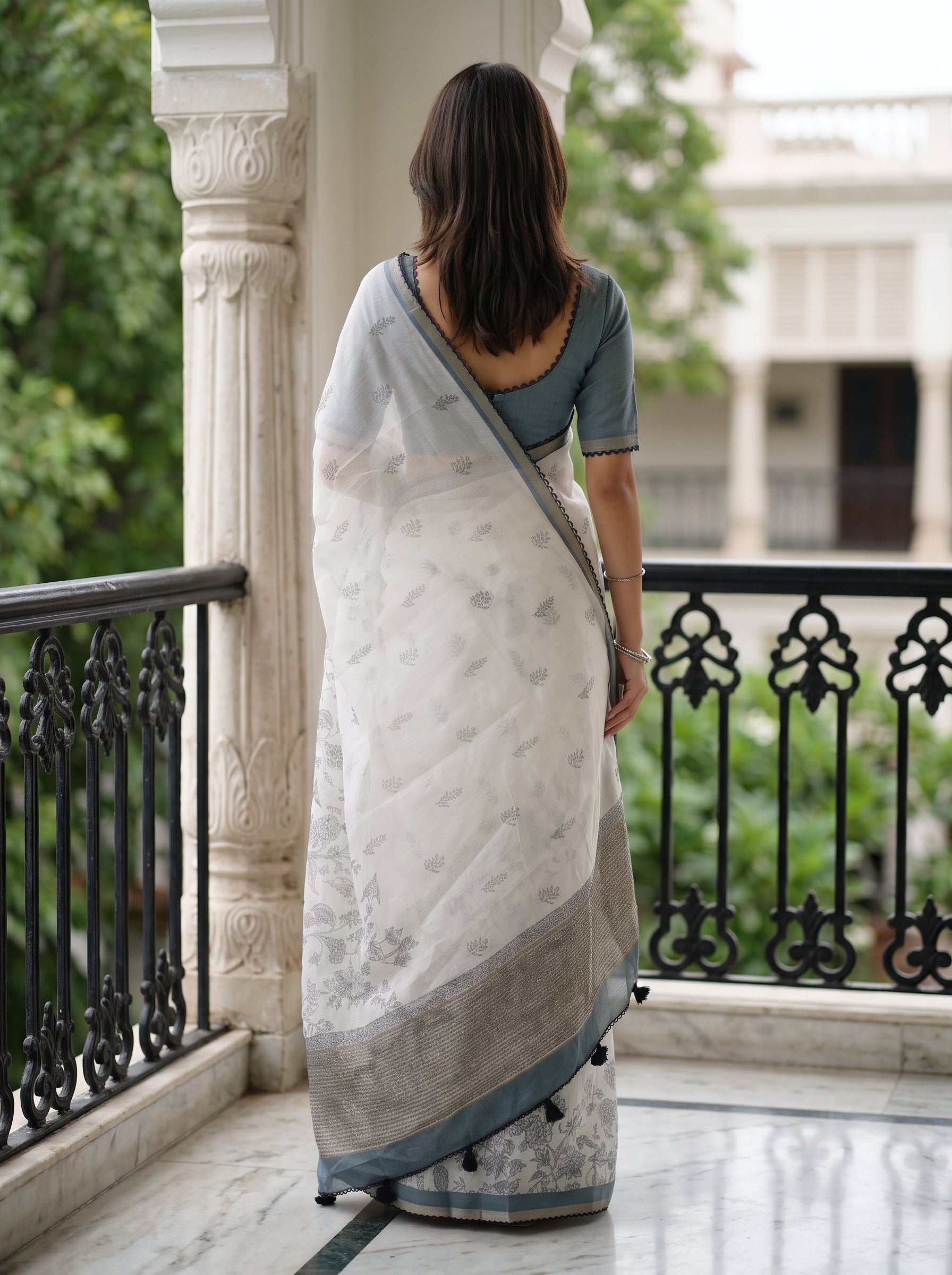 A woman viewed from behind wearing a white and gray saree with a dusty blue blouse, standing on a marble balcony with a black railing.