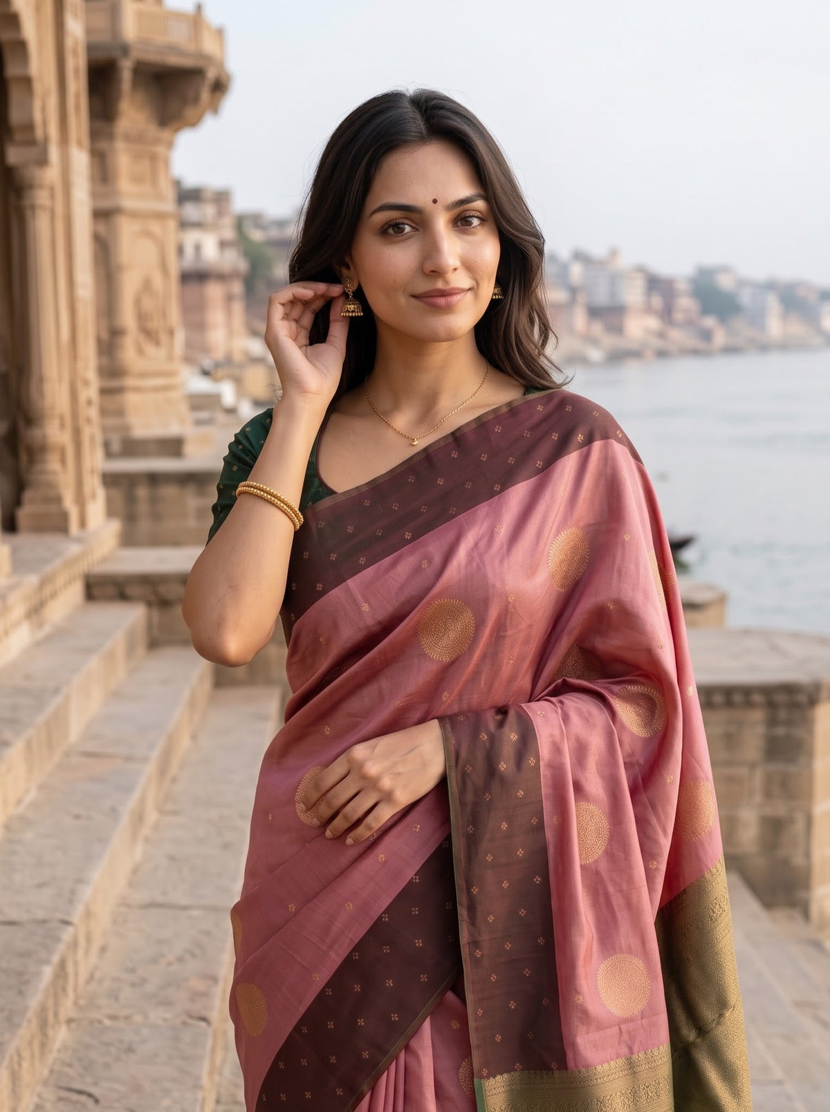 A woman wearing a dusty pink and dark brown silk saree with gold motifs, standing on stone steps with a river and cityscape in the background.