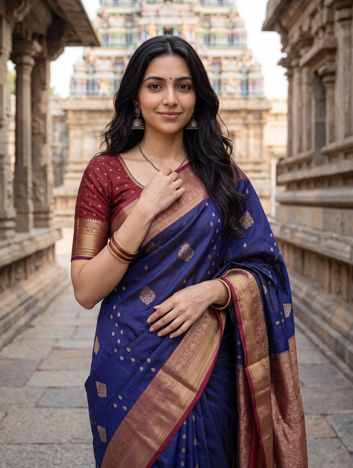A woman wearing a royal blue silk saree with gold zari motifs and a deep red blouse, standing in a stone corridor with a temple gopuram in the background.