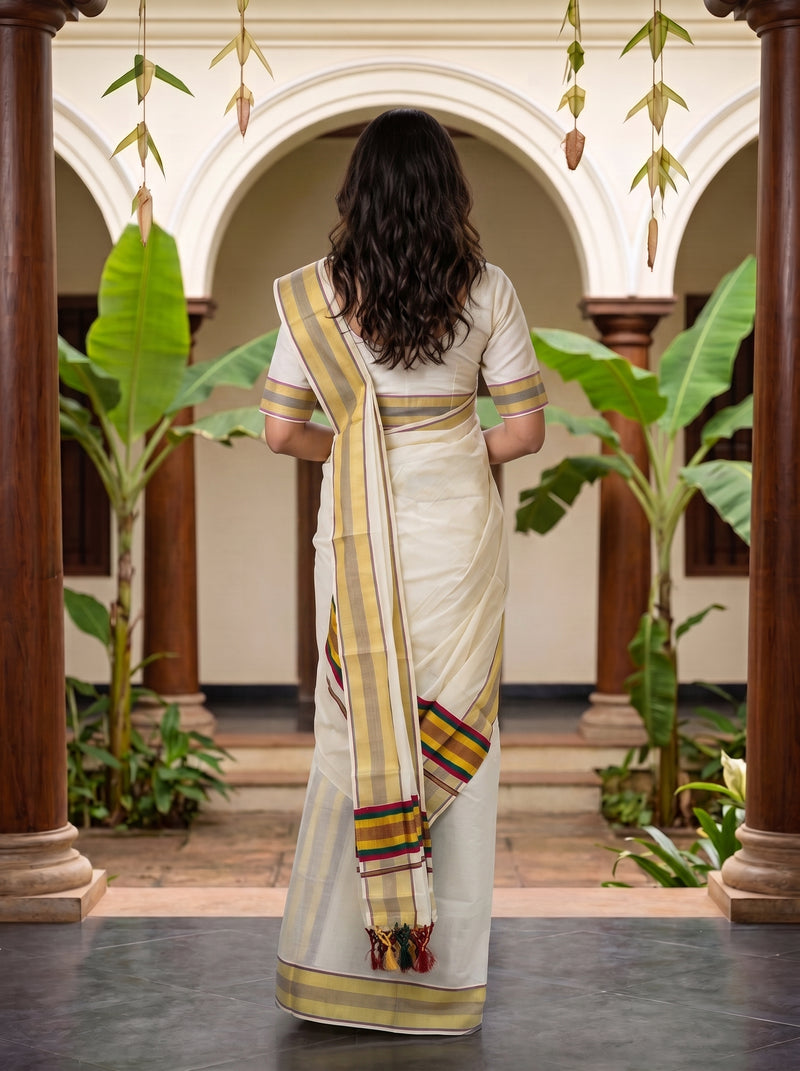 A woman viewed from behind wearing a cream-colored saree with yellow, red, and green striped borders, standing in a courtyard with arches and potted plants.