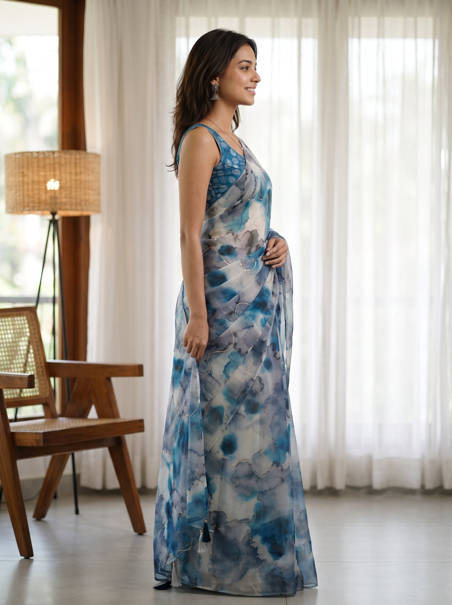 A woman wearing a sleeveless blue and white abstract-print maxi dress, standing in profile in a bright room with sheer white curtains, a wooden chair, and a floor lamp.