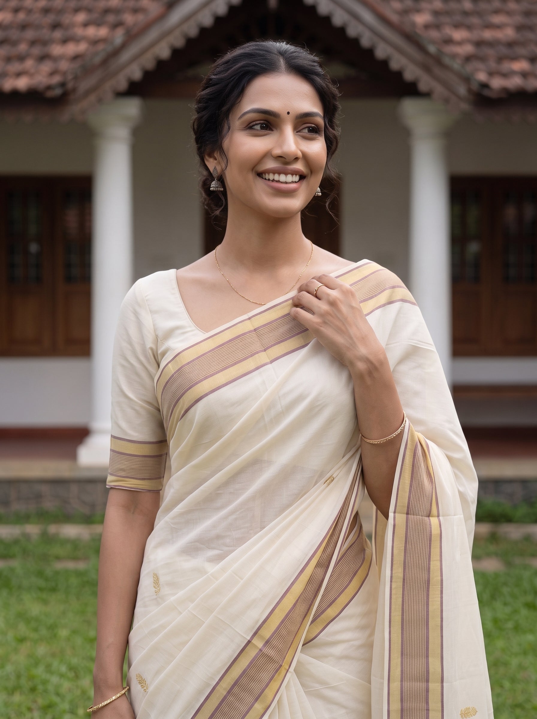 A woman wearing a cream-colored cotton saree with beige and gold stripes, standing outdoors in front of a traditional house with white pillars and a tiled roof.