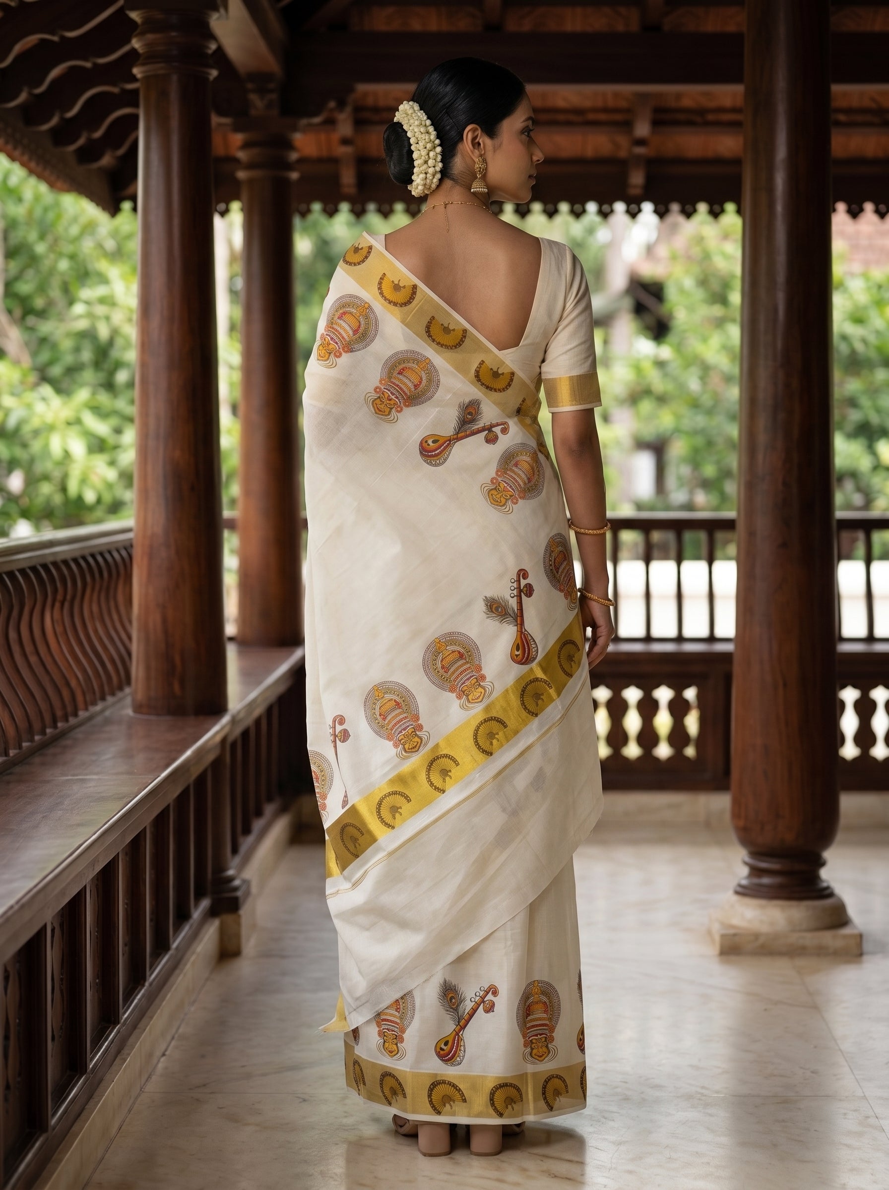 A woman wearing a traditional white saree with gold and brown motifs, standing on a wooden porch.
