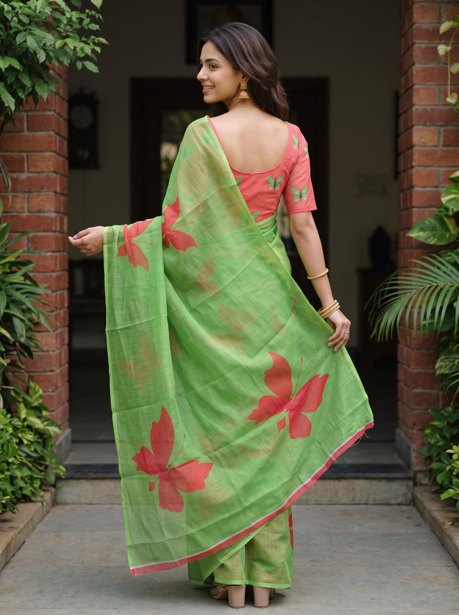 A woman wearing a light green saree with large red floral prints and a coral pink blouse, standing in a doorway between brick pillars and green plants.