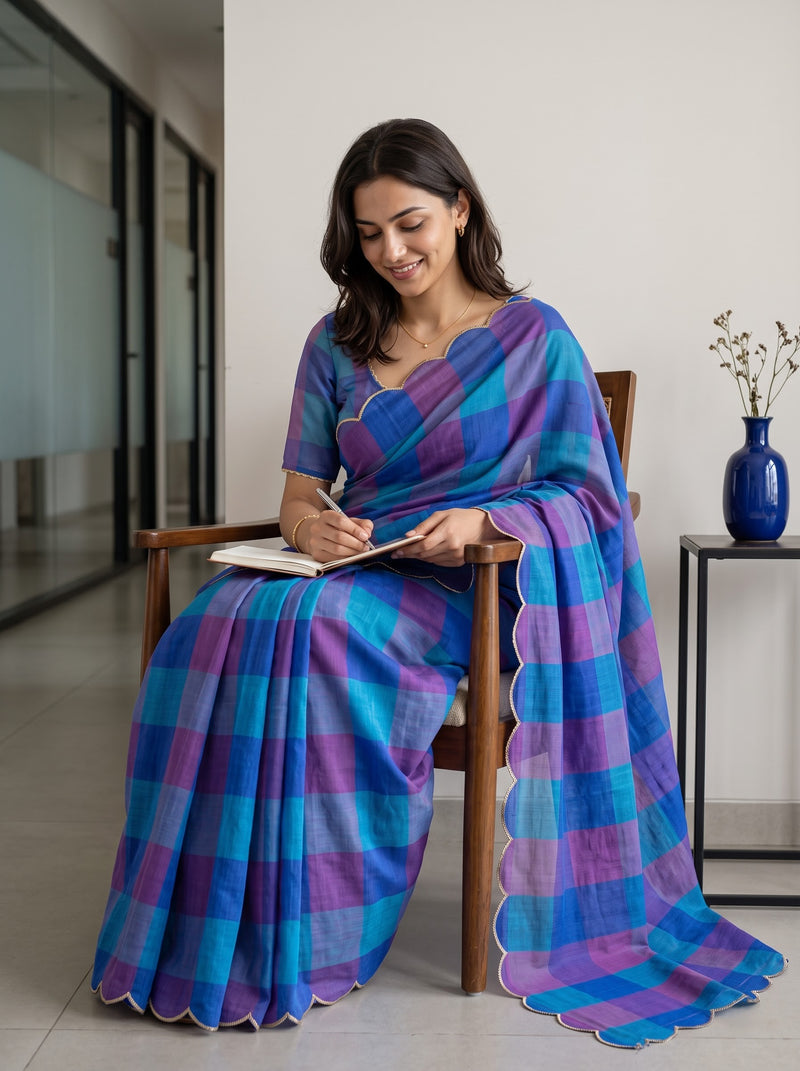 A woman sits on a wooden chair in a modern indoor setting, wearing a blue and purple checkered saree and writing in a notebook.