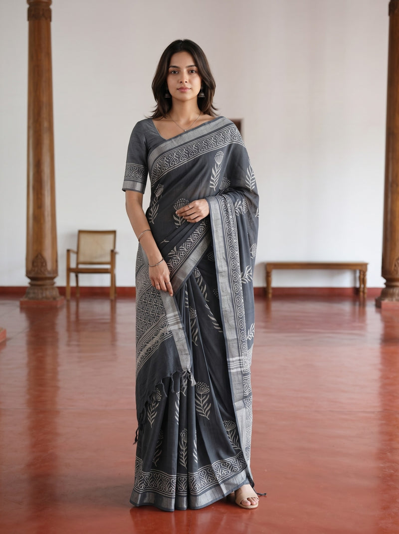 A woman modeling a dark gray and silver printed saree with a matching short-sleeve blouse in a spacious room with wooden pillars and a polished red-brown floor.