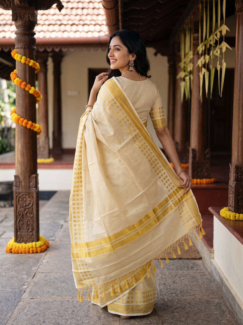 A woman wearing a light beige and yellow saree stands in a traditional courtyard, holding out the saree drape to show its border and tassel details.
