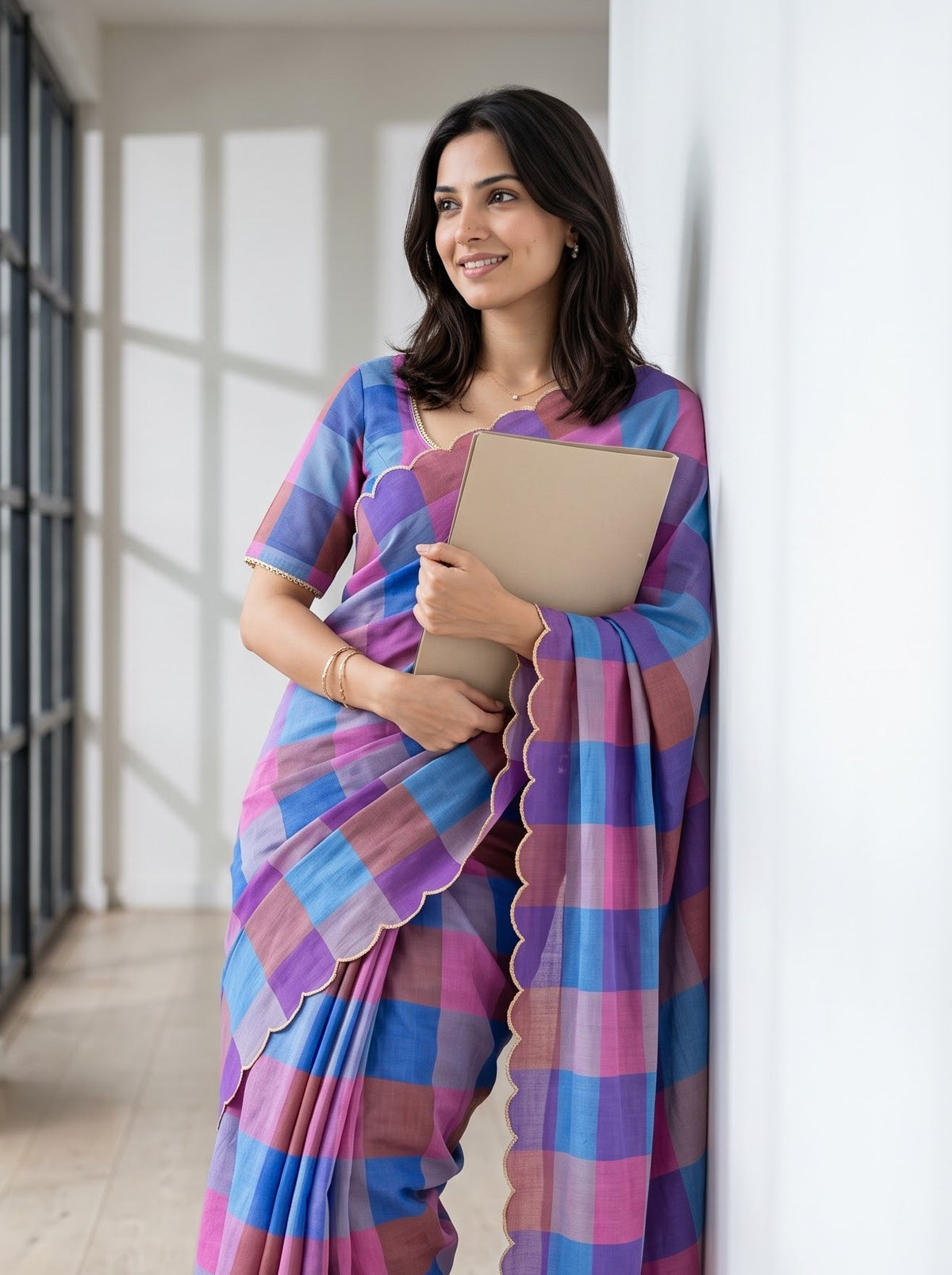 A woman wearing a colorful checkered saree with purple, blue, and pink tones, holding a beige folder against a bright indoor background.