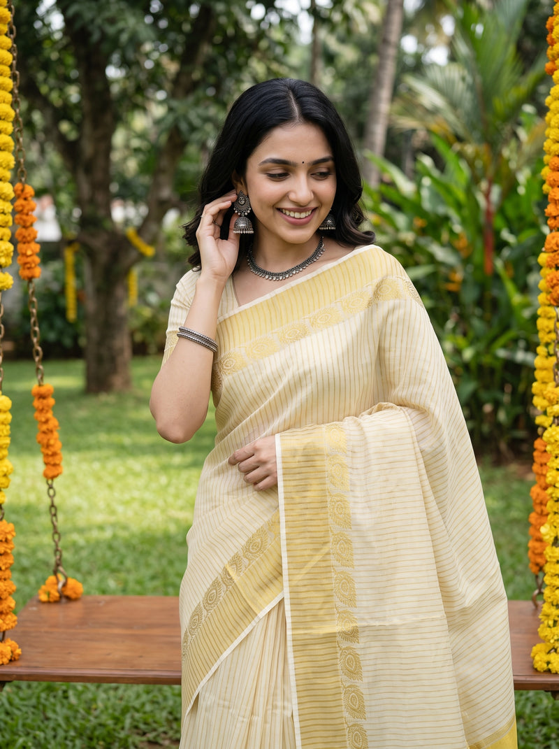 A woman wearing a light cream-colored saree with subtle vertical stripes and a patterned border, standing outdoors on a wooden swing surrounded by green foliage and yellow flower garlands.