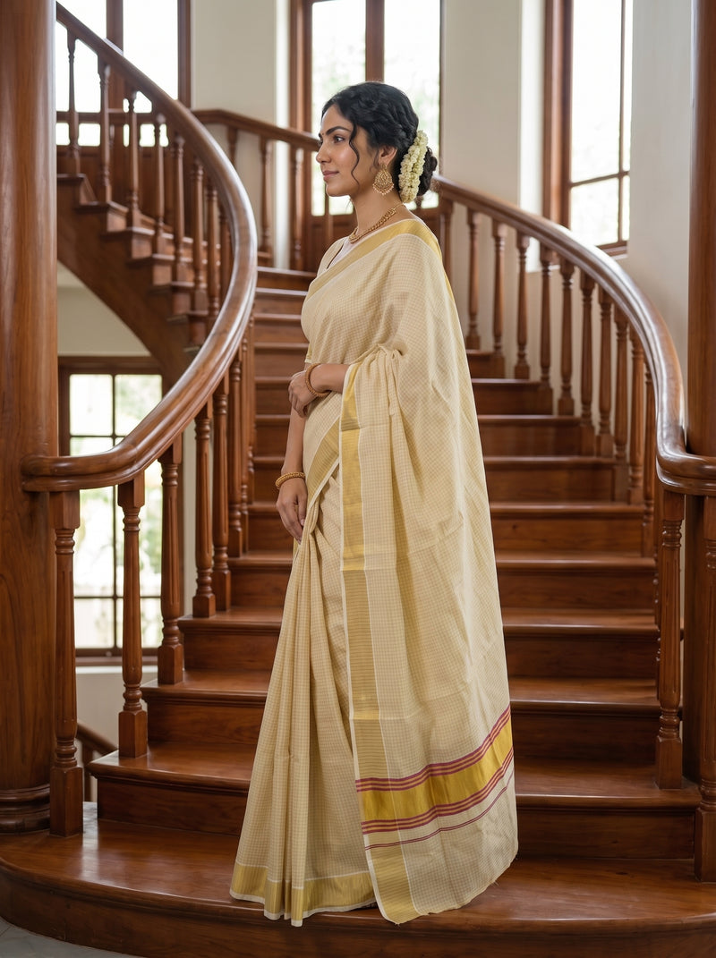 A woman wearing a cream-colored silk saree with yellow and red border stripes stands on a wooden staircase in a traditional Indian setting.