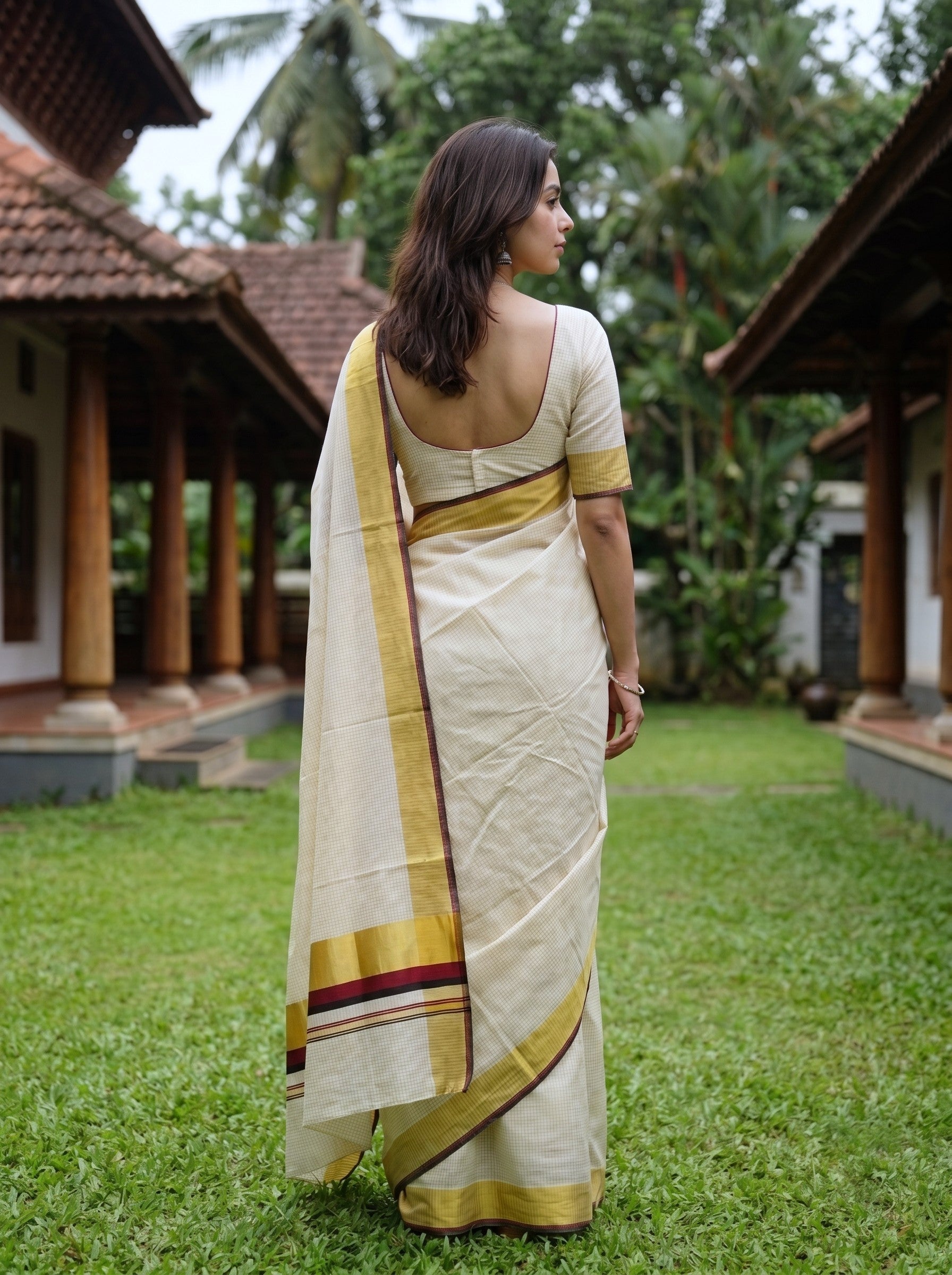 A woman wearing a cream-colored cotton saree with gold and maroon borders, standing in a garden courtyard with traditional houses and green grass in the background.