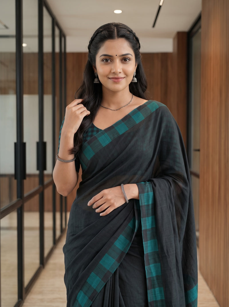 A woman wearing a dark charcoal gray saree with a teal green and black checkered border, standing in a modern office hallway with glass walls and wood paneling.