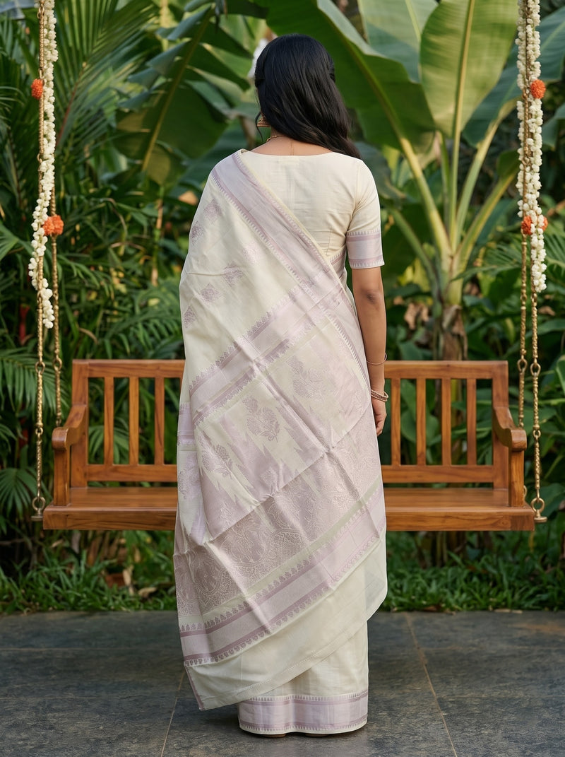 A woman viewed from behind wearing a light cream or off-white saree with a lavender-pink border and pallu, standing in front of a wooden swing in a lush garden setting.