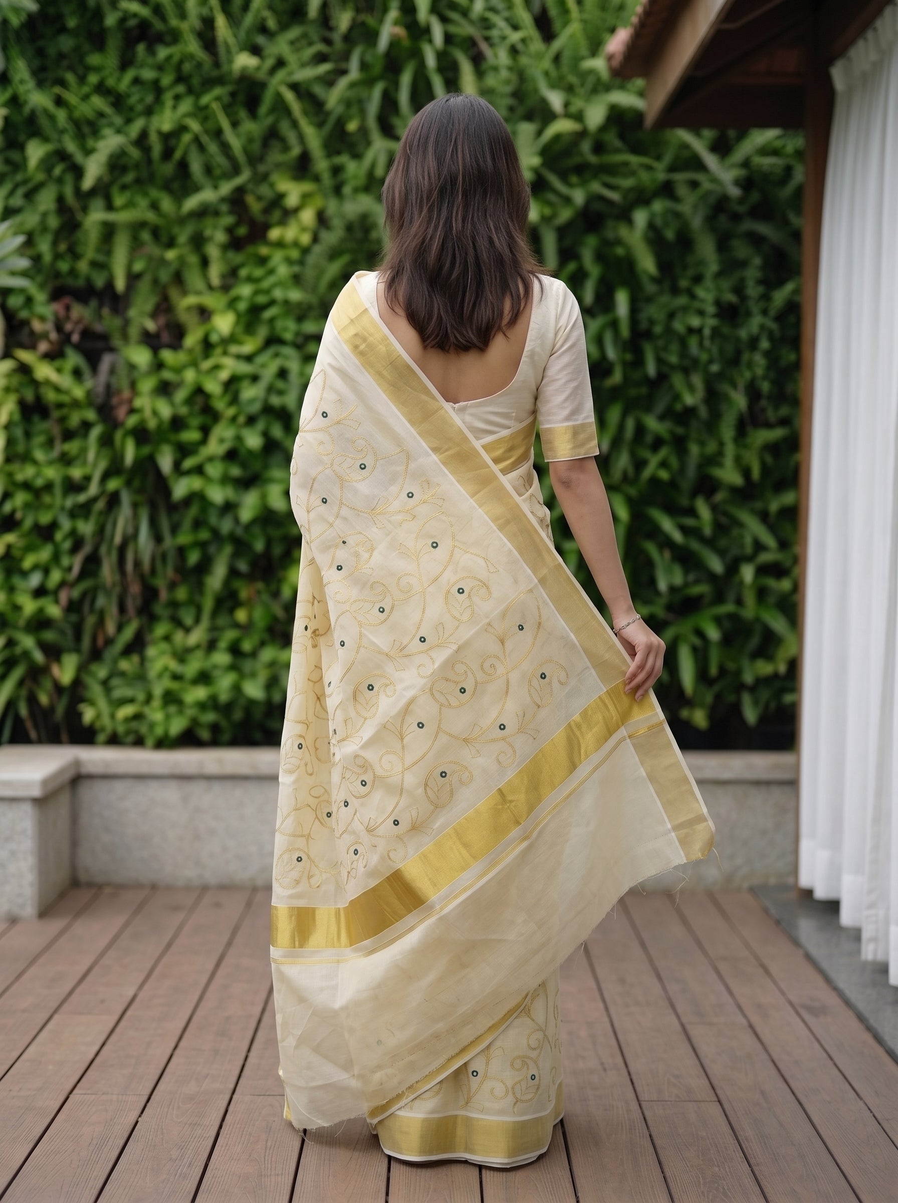 A woman viewed from behind wearing a cream-colored saree with gold borders and motifs, standing on a wooden deck in front of a green leafy wall.