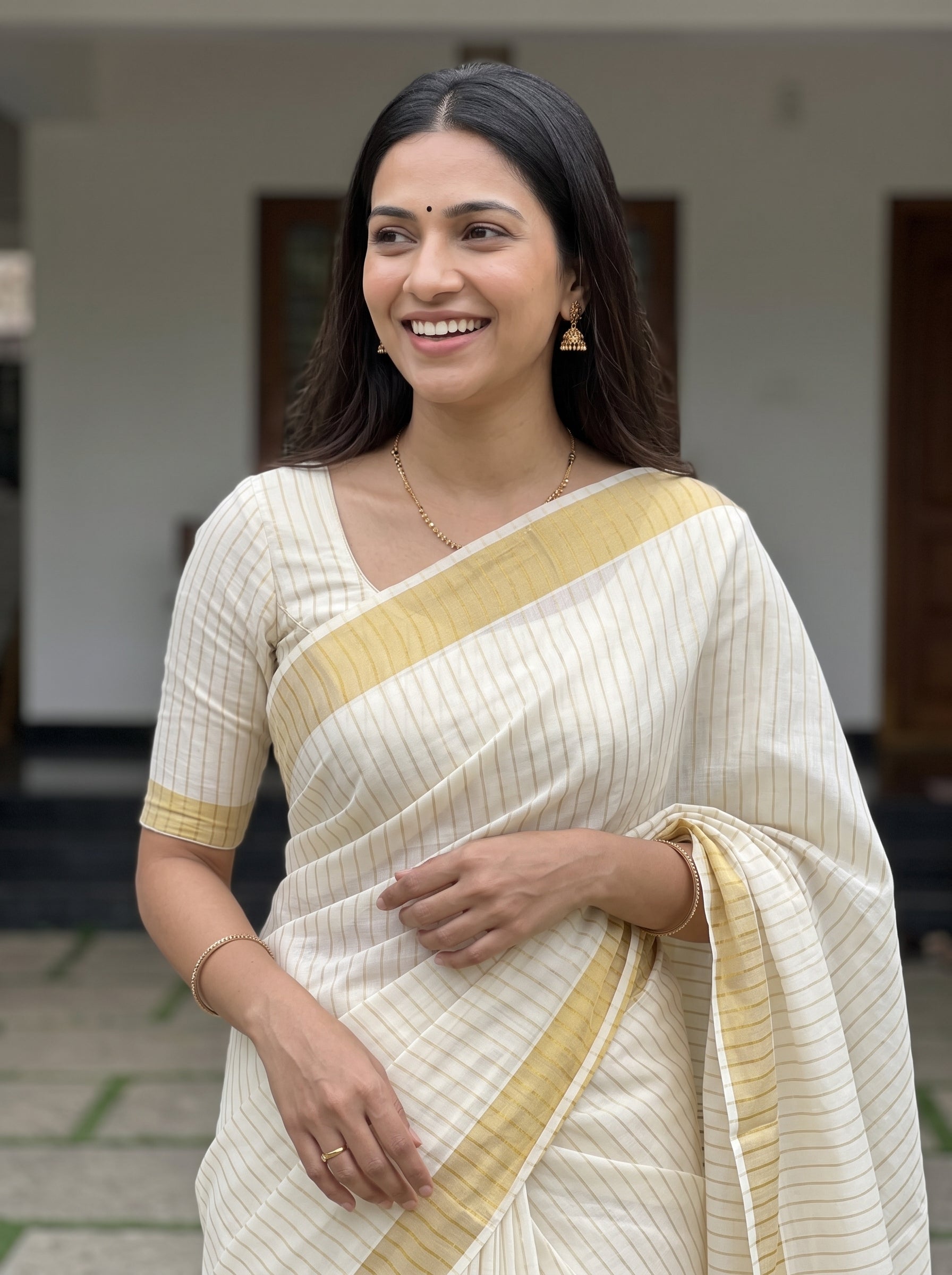 A woman wearing a cream-colored sari with thin vertical stripes and a mustard yellow border, standing in an outdoor setting.