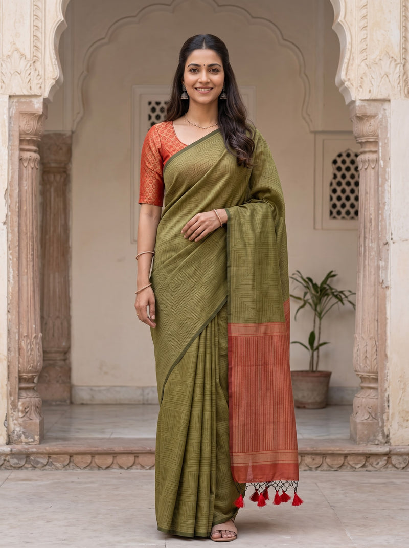 A woman modeling a sage green silk saree with a rust-orange border and matching blouse, standing in a traditional courtyard with arches.