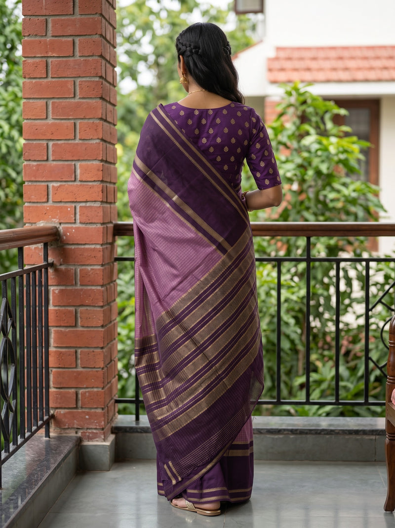 A woman viewed from behind wearing a purple saree with gold patterns and a matching purple blouse, standing on a balcony with a brick pillar and green garden background.
