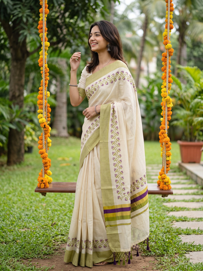 A woman wearing a cream and olive green saree with floral embroidery and a purple accent stripe, standing next to a wooden swing decorated with orange marigold garlands in a garden setting.