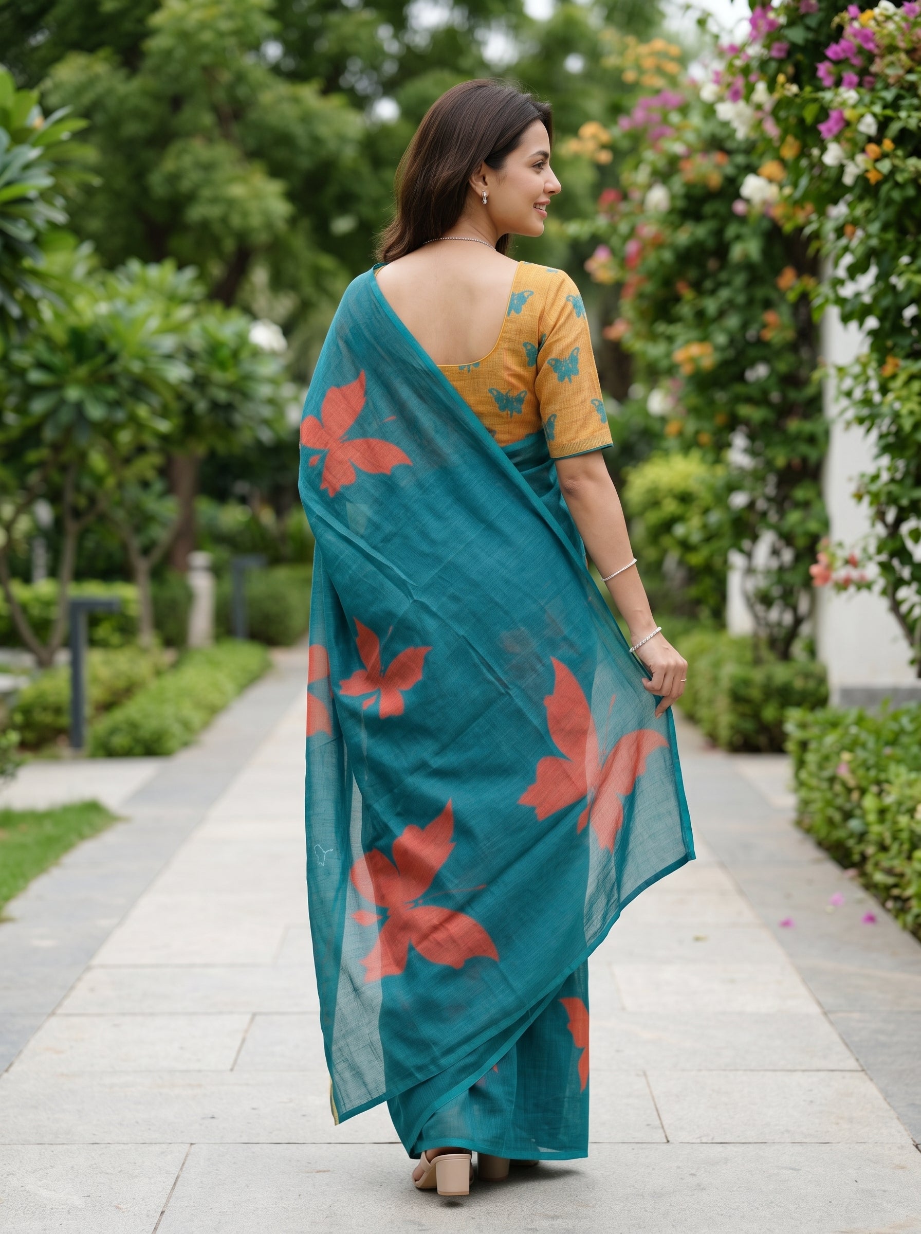 A woman wearing a teal blue saree with red floral prints and a mustard yellow printed blouse, standing on a stone pathway in a garden.