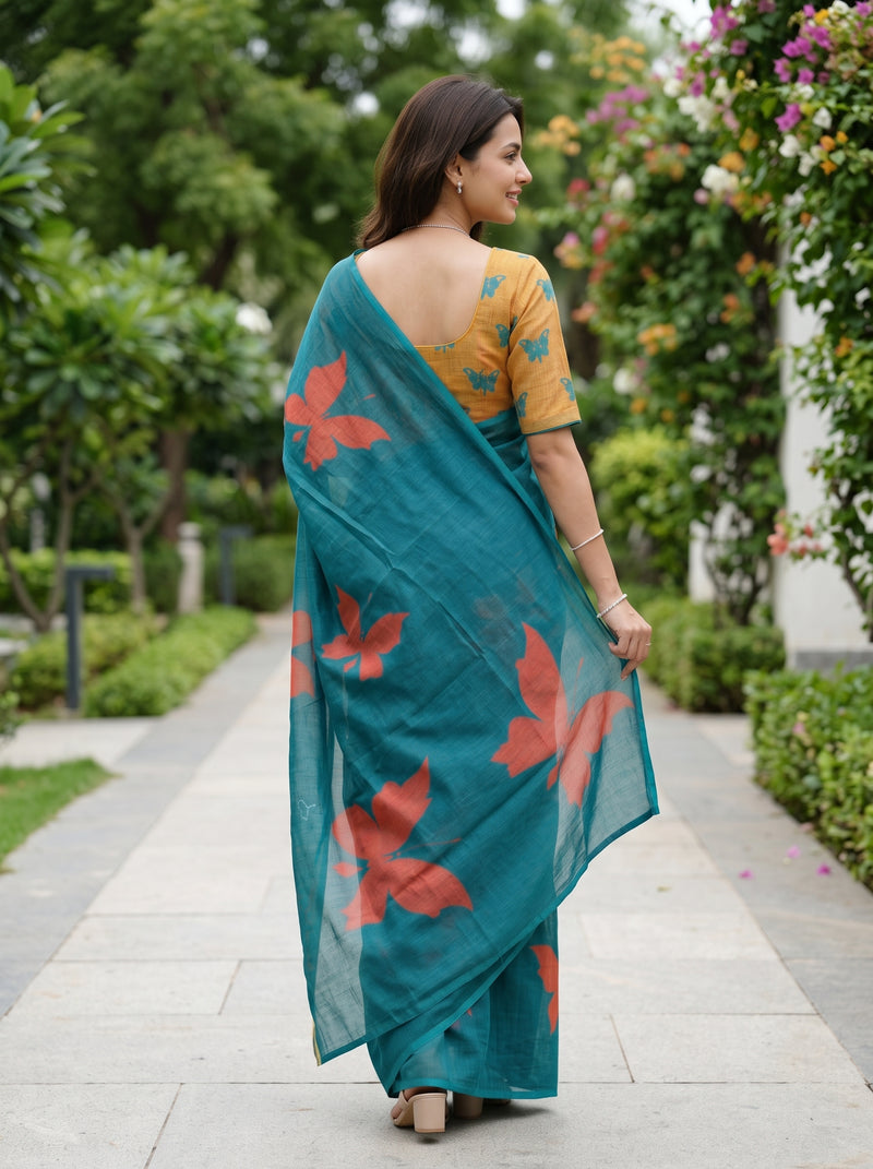 A woman wearing a teal blue saree with red floral prints and a mustard yellow printed blouse, standing on a stone pathway in a garden.