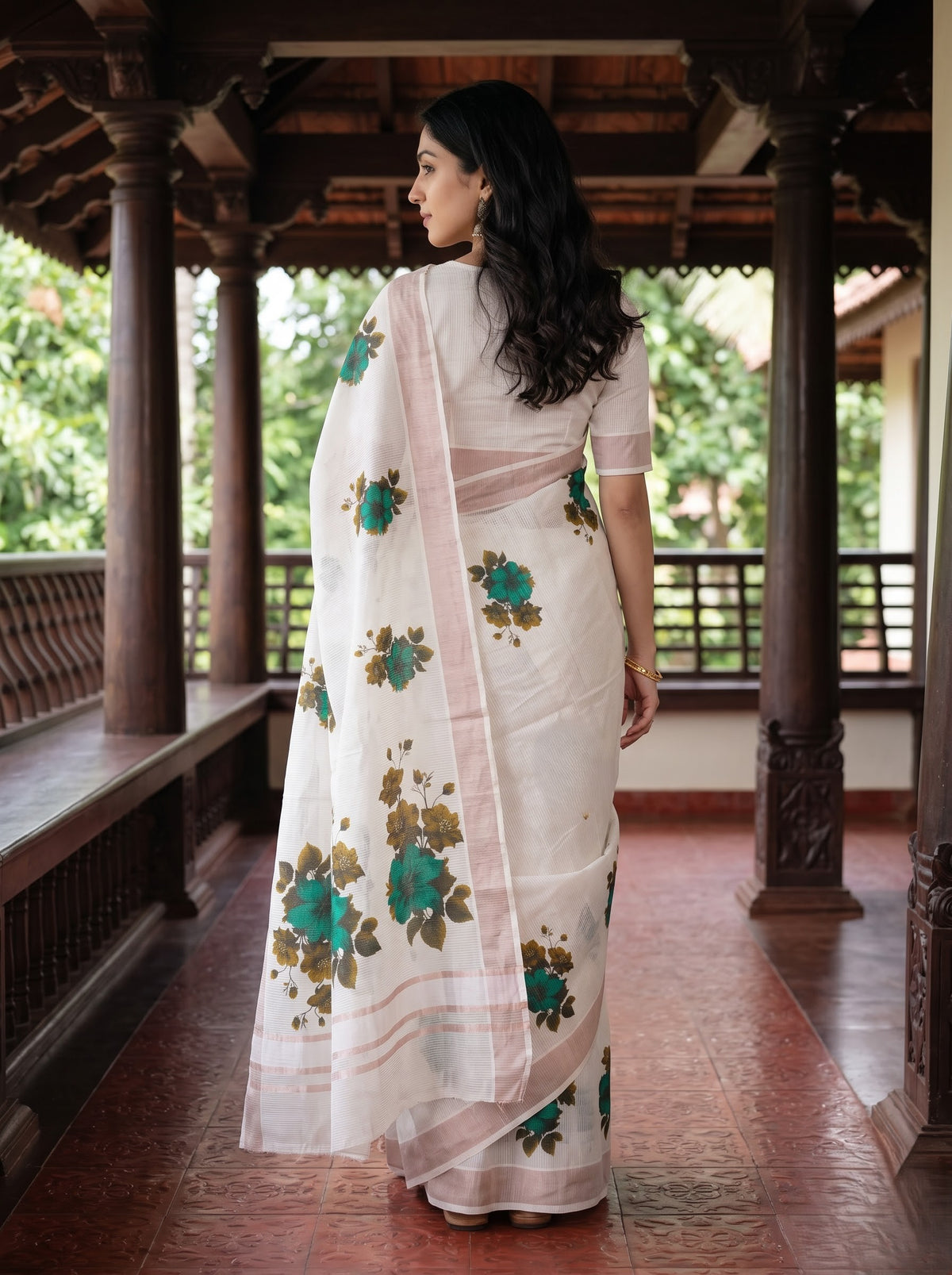 A woman wearing a white saree with green and gold floral patterns and a pink border, standing in a traditional wooden corridor.