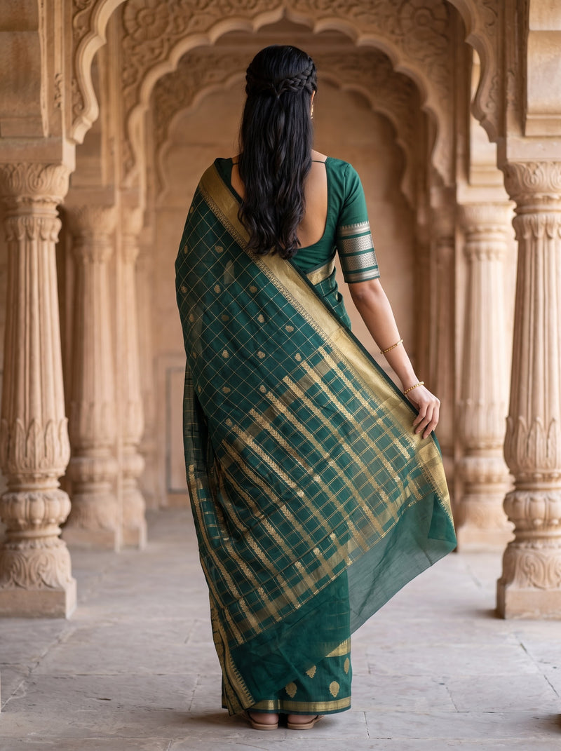 A woman viewed from behind wearing a dark green saree with gold stripes and a matching green-and-gold blouse, standing in a stone corridor with arches.