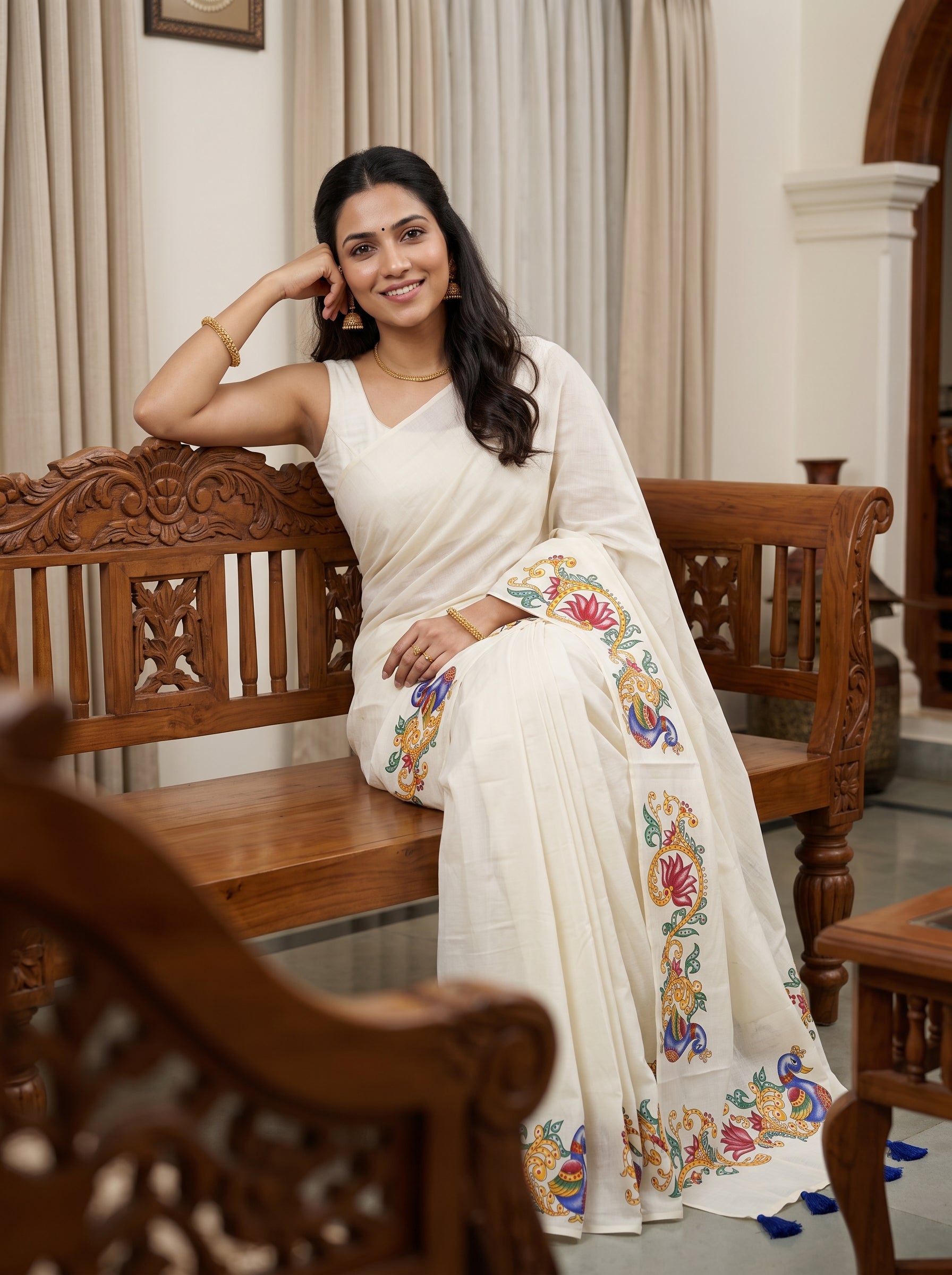 A woman wearing a white saree with colorful floral embroidery sits on a carved wooden bench in an indoor setting with beige curtains.